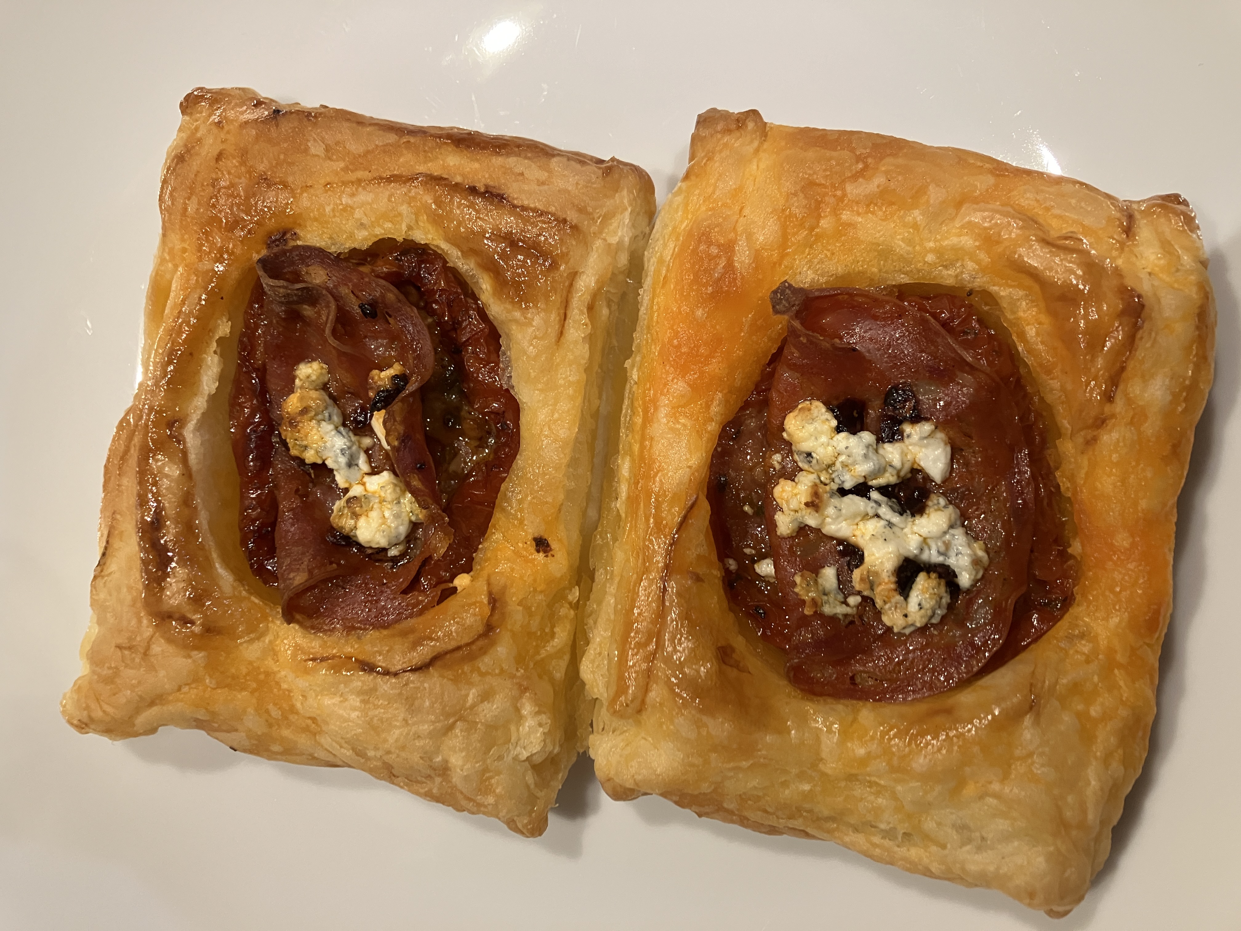 two pastry rectangles with sundried tomatoes, chorizo, and golden brown goats cheese in the middle, on a white plate two pastry rectangles with sundried tomatoes, chorizo, and golden brown goats cheese in the middle, on a white plate