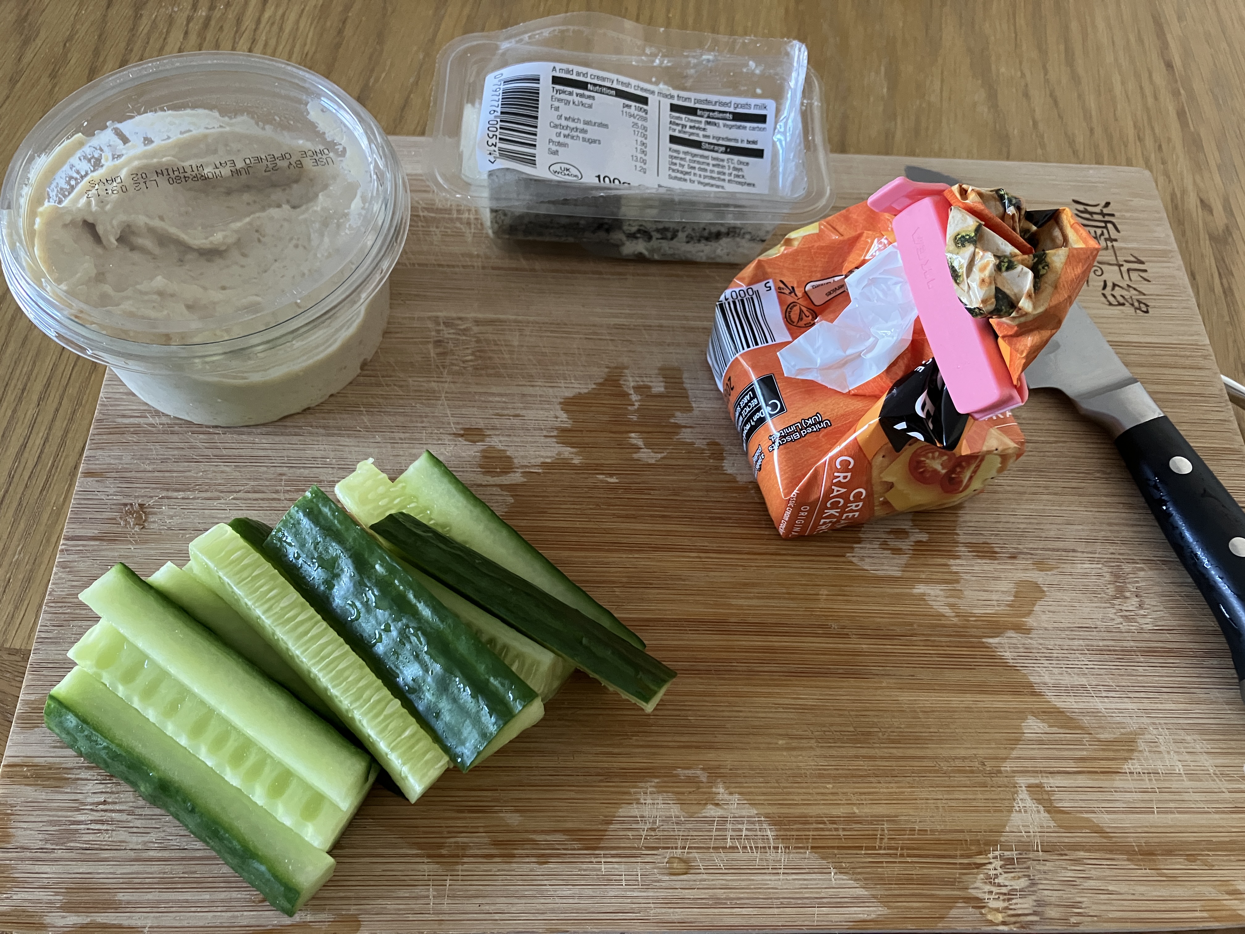 some food on a wooden chopping board: cucumber sticks, a pot of hummus, a pot of goats cheese, and a small pack of jacob's cream crackers some food on a wooden chopping board: cucumber sticks, a pot of hummus, a pot of goats cheese, and a small pack of jacob's cream crackers